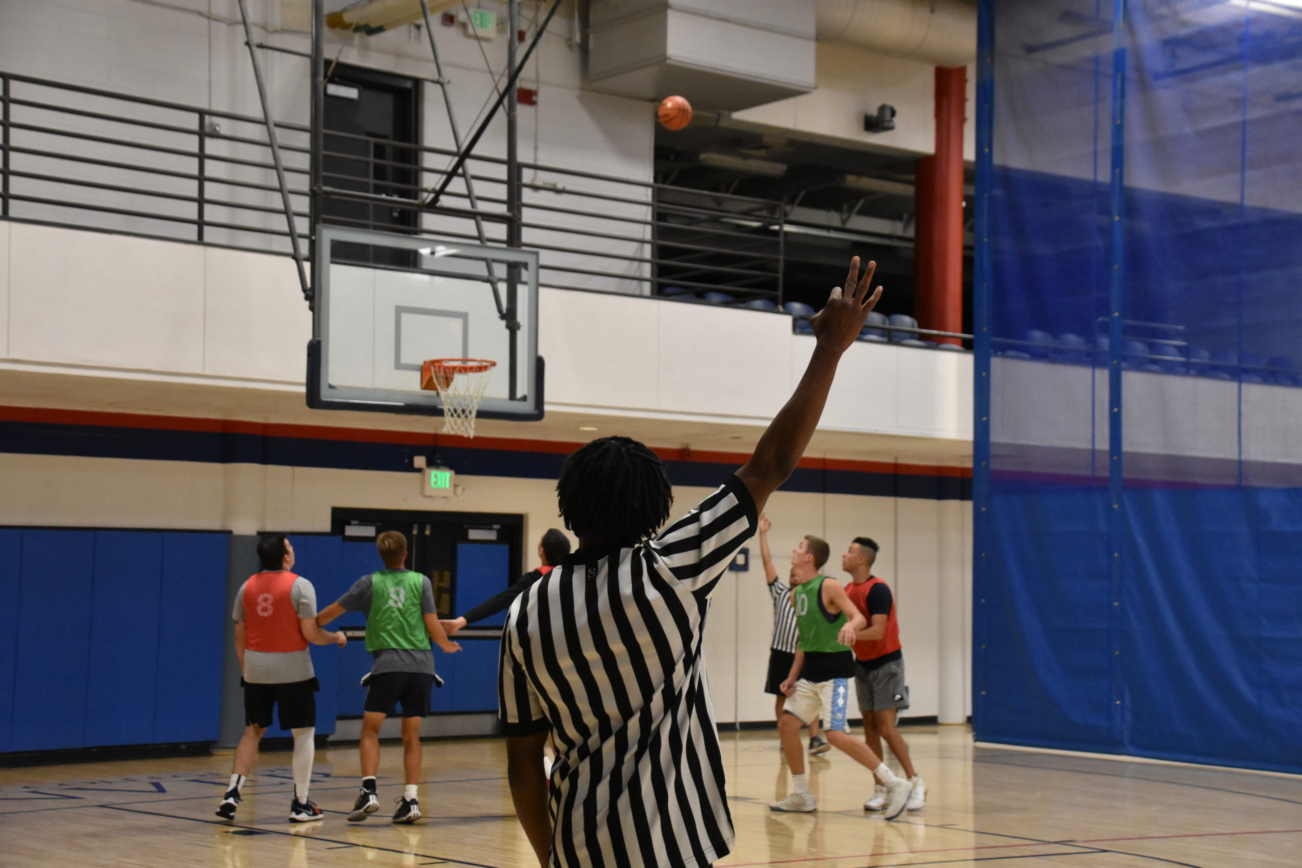 A basketball game is underway in an indoor gymnasium, with players wearing colored practice jerseys and a referee in a black-and-white striped shirt raising an arm to signal a call. A basketball is visible in the air near the hoop, with the court, bleachers, and gym equipment in the background.