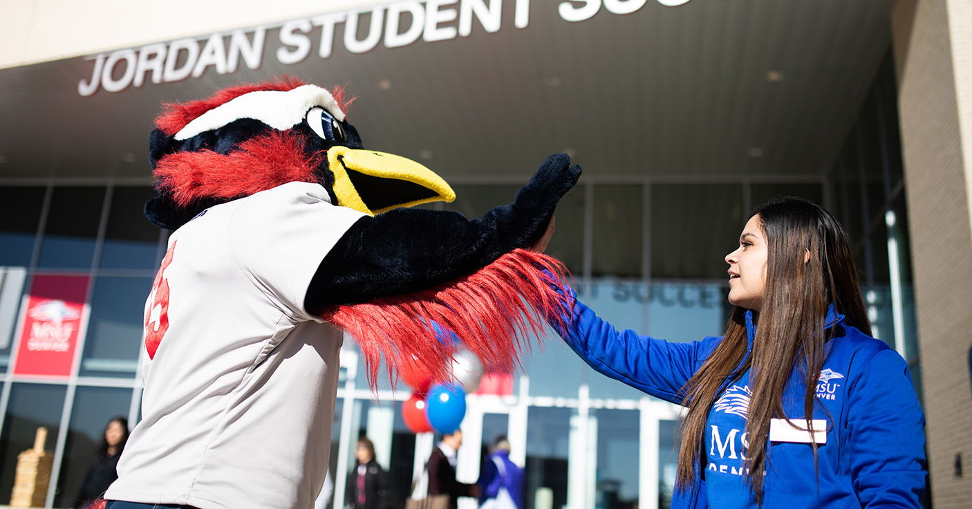 Rowdy the Roadrunner gives a high-five to a student outside a building entrance. The scene takes place in front of a modern glass building with signage above the entrance, while other people and colorful balloons are visible in the background.
