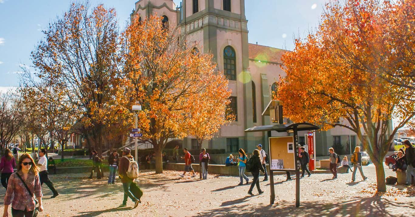 A sunny outdoor campus scene with people walking along a pathway lined with trees displaying orange autumn foliage. A large building with a tower is visible in the background, and students move through the plaza area near a campus map sign.