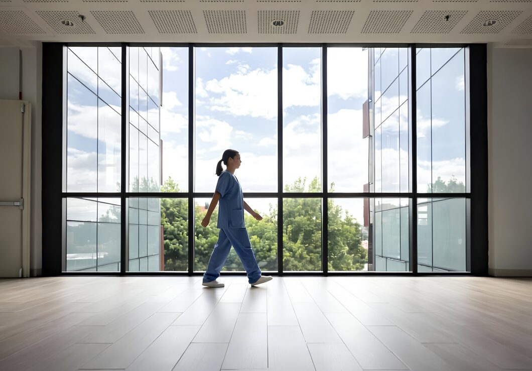 A person wearing blue medical scrubs walks through a bright, modern interior hallway with large floor-to-ceiling windows. Natural light fills the open space, with trees and buildings visible outside through the glass. 