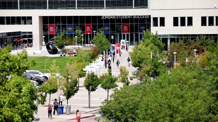A wide view of the Jordan Student Success building and surrounding campus plaza on a sunny day. Tree-lined walkways lead to the building entrance, where people walk, gather, and sit outdoors. A large sculpture, landscaped green spaces, and campus banners are visible in the foreground.