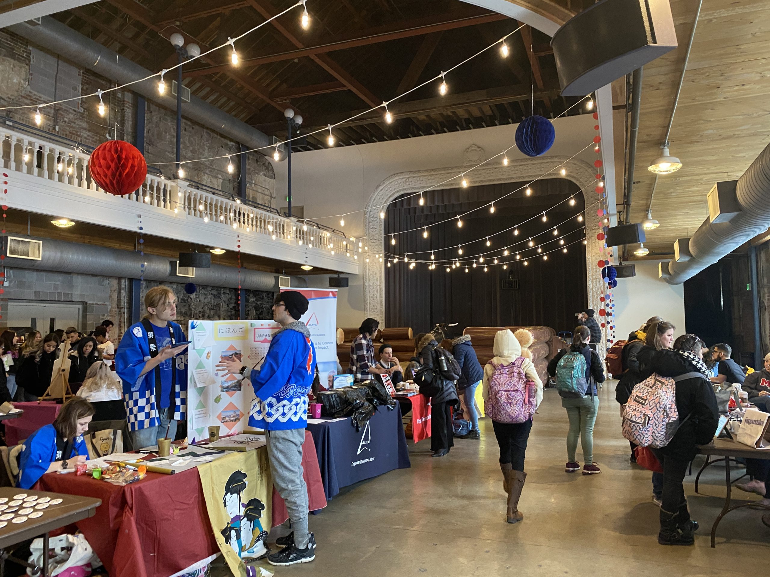 A large indoor student organization fair takes place in a spacious hall with high ceilings, exposed beams, and string lights. Attendees browse information tables, talk with student organization representatives, and walk through the event. Booths with posters and displays line both sides of the room, while a stage is visible at the far end.