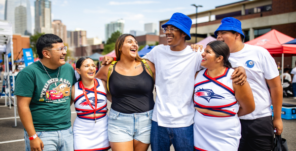 Five people stand together outdoors at a campus event with their arms around one another, smiling and laughing. Two wear cheer uniforms, two wear blue bucket hats, and one wears a green T-shirt. Colorful event tents and a city skyline are visible in the background.