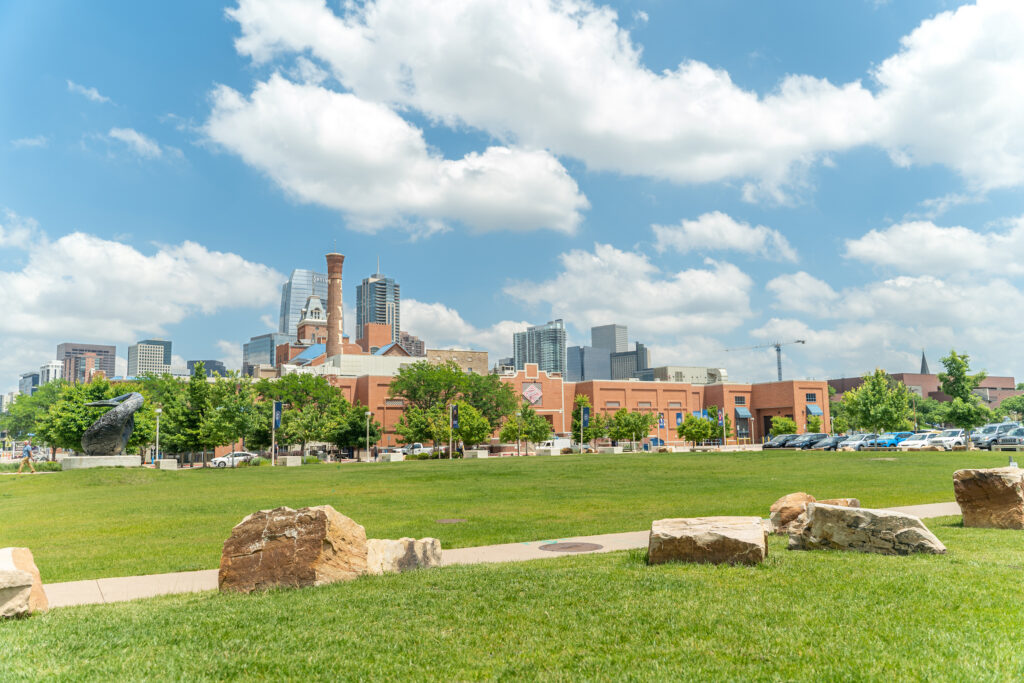 Denver skyline viewed from a grassy area, with large rocks in the foreground, trees and the Tivoli building in the middle ground, and downtown high-rise buildings beneath a bright blue sky with clouds.