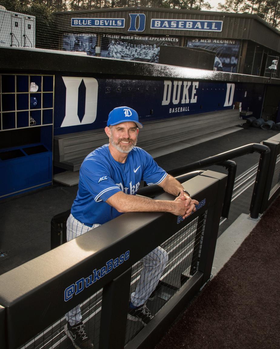 Duke baseball Coach Chris Pollard, wearing a Duke blue baseball uniform is standing in the dugout while smiling