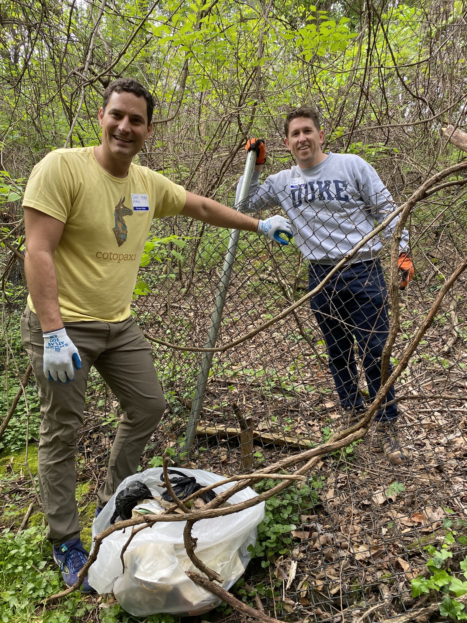 Two men smile while participating in an outdoor cleanup project. One leans on a chain-link fence holding work gloves, while the other stands beside a large plastic bag filled with debris and vines. They are surrounded by dense woods and undergrowth.
