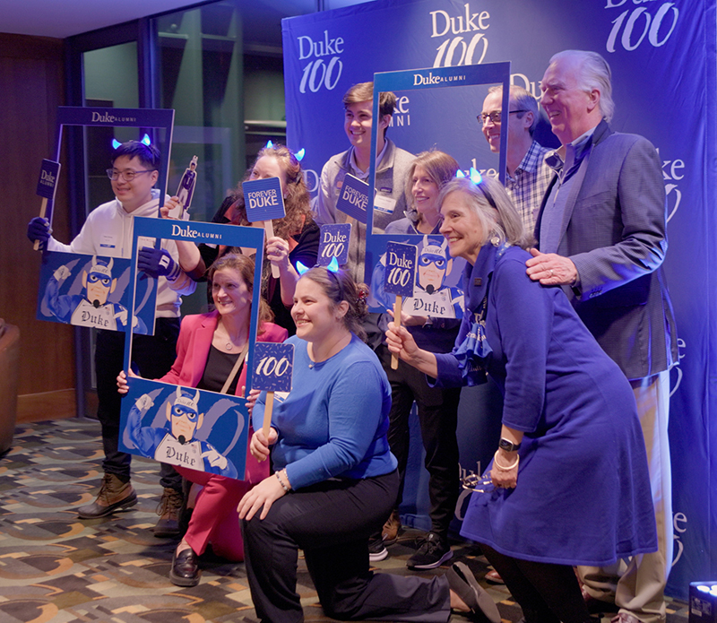 A group of Duke alumni and supporters pose together at a Duke 100 event in Boston. They are smiling and holding Duke-themed props, including “Forever Duke” signs, cutouts of the Blue Devil mascot, and Duke Alumni photo frames. Some participants wear blue glowing headbands shaped like horns. A blue Duke 100 step-and-repeat banner serves as the backdrop.