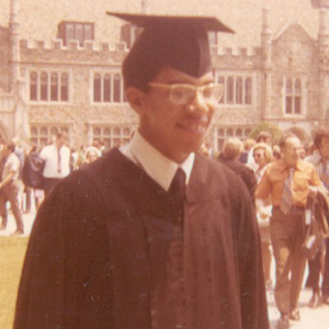 A young man in graduation attire smiles while standing outdoors at Duke University. He wears a cap, gown, and glasses, with Duke’s historic stone architecture and a crowd of people in the background.