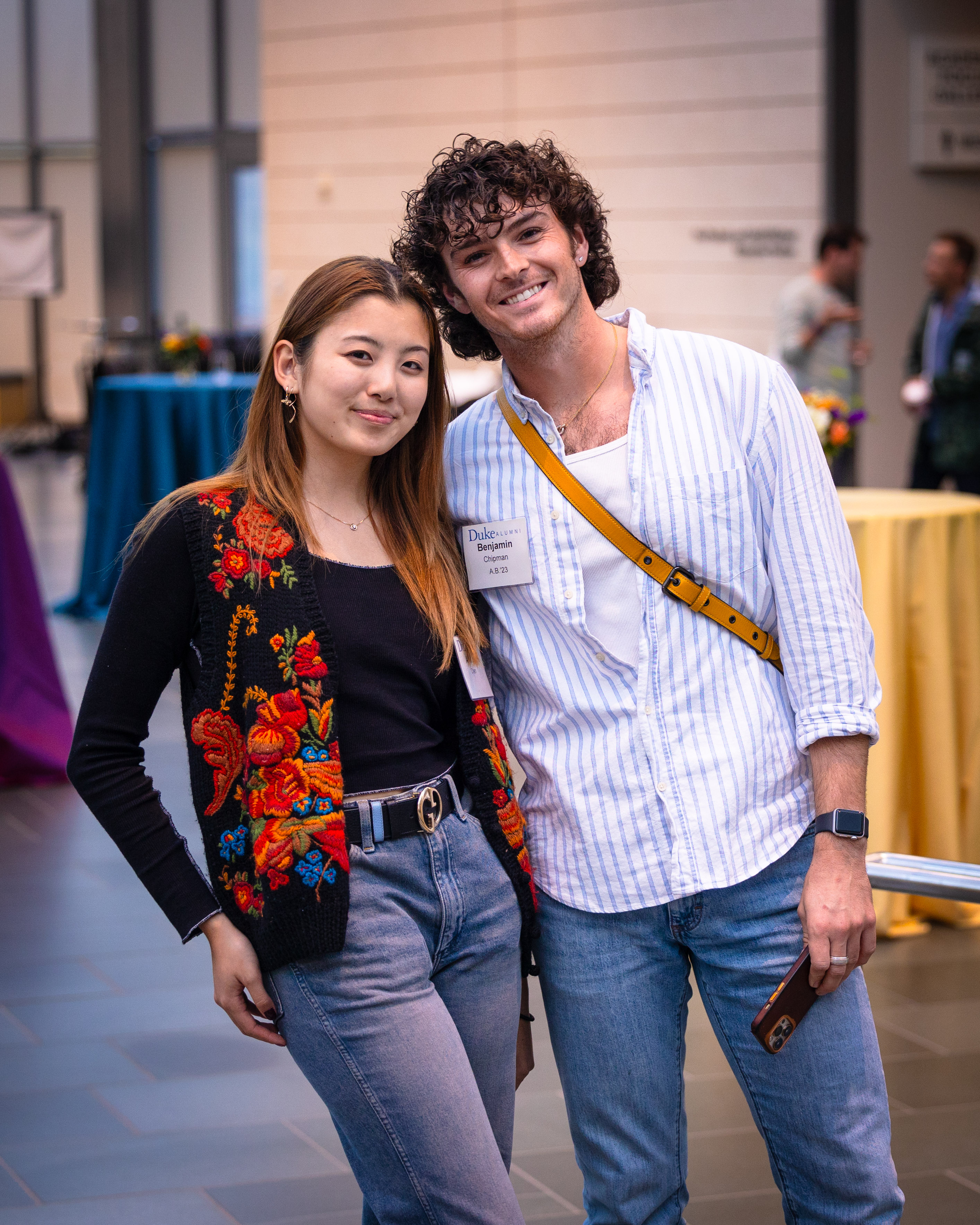 Two young adults smile while standing together at an indoor Duke alumni event. One wears a black floral vest and jeans, and the other wears a striped shirt with a name badge and a yellow crossbody strap. High-top tables and other event guests appear blurred in the background.
