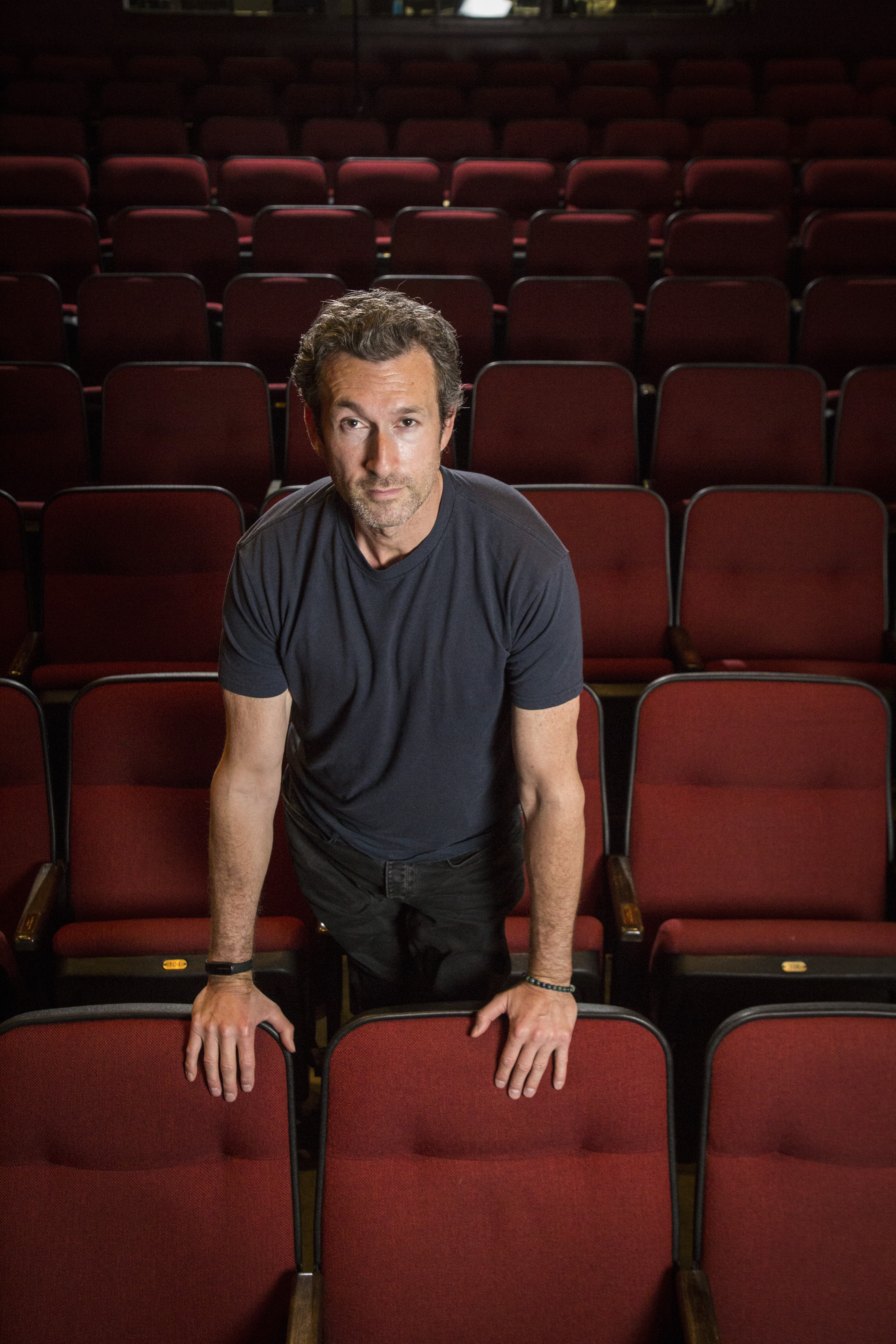 A person stands in a dimly lit theater surrounded by empty red seats, leaning forward on a chair and looking confidently toward the camera.
