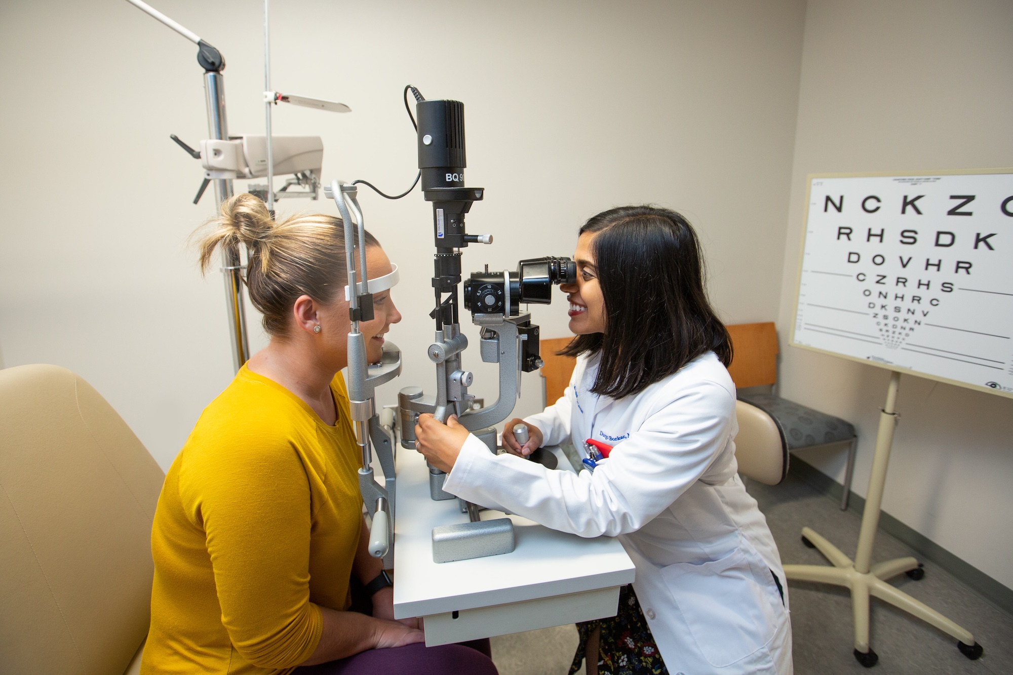 A healthcare professional in a white lab coat examines a patient’s eyes using a slit-lamp microscope in a clinic room. The patient sits still with their chin on the machine’s support, while an eye chart stands in the background.