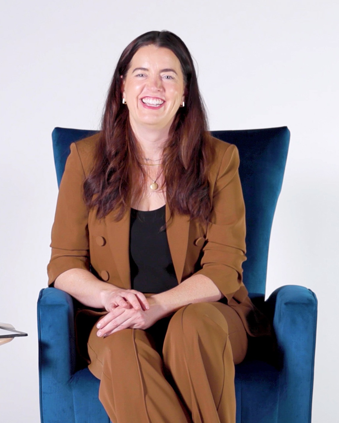 Kate Bowler sits in a blue velvet armchair, smiling warmly at the camera. She’s wearing a brown pantsuit over a black top, with simple jewelry. Her hands rest casually in her lap, and the background is a clean white studio setting.