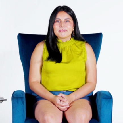 Victoria Pinedo sits in a blue velvet armchair, smiling warmly at the camera. She’s wearing a brown pantsuit over a black top, with simple jewelry. Her hands rest casually in her lap, and the background is a clean white studio setting.