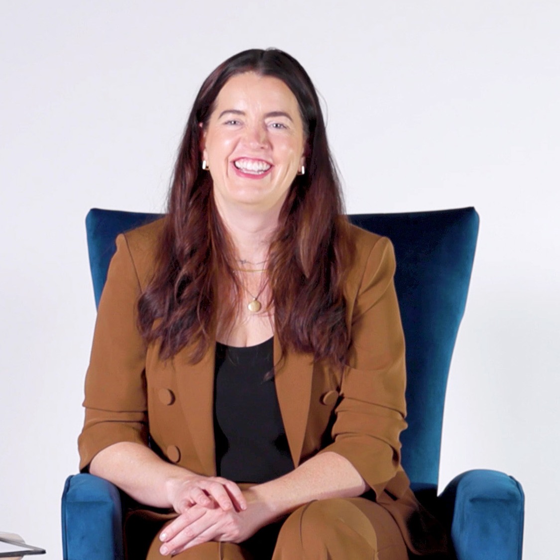 Kate Bowler sits in a blue velvet armchair, smiling warmly at the camera. She’s wearing a brown pantsuit over a black top, with simple jewelry. Her hands rest casually in her lap, and the background is a clean white studio setting.