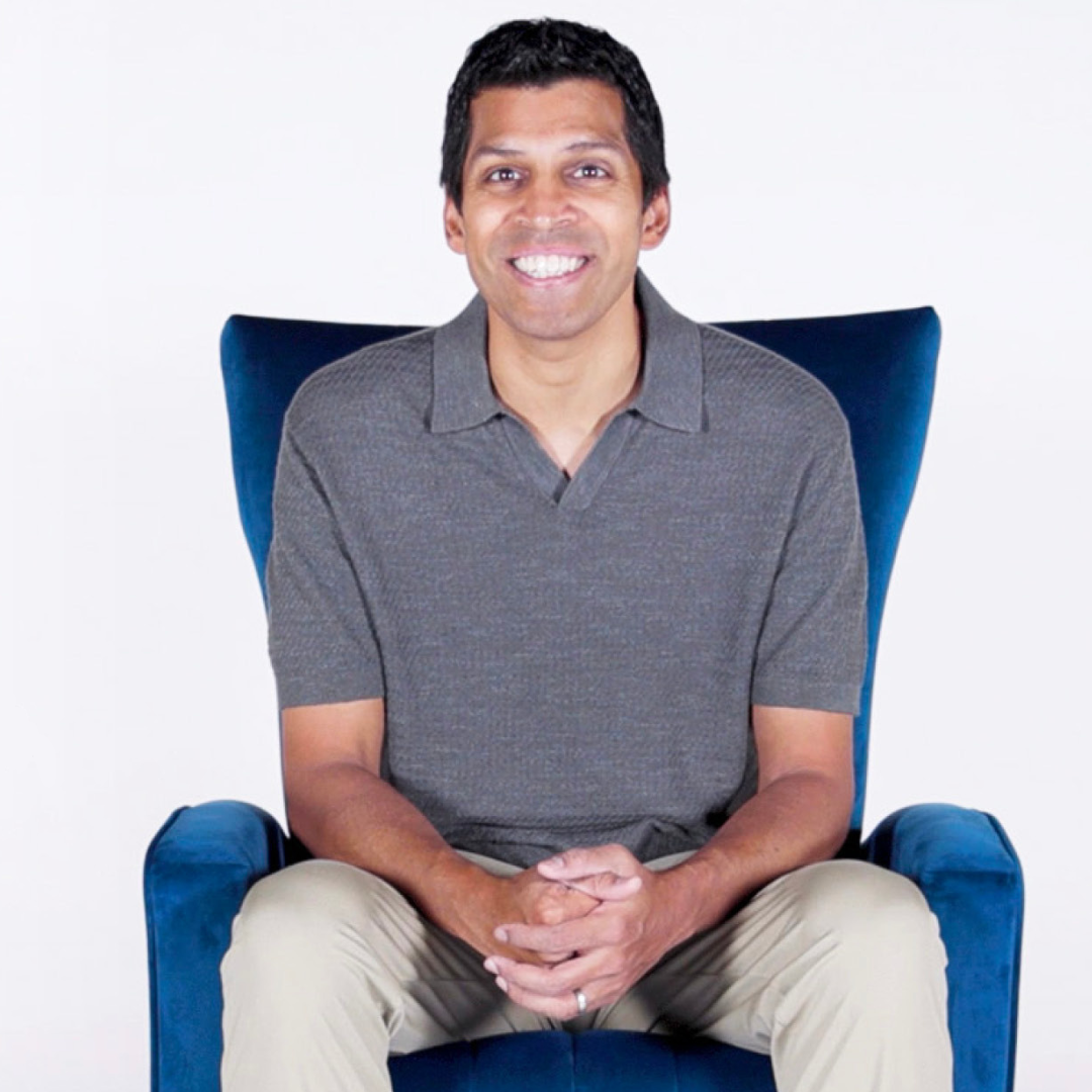 Ronnie Chatterji sits smiling in a deep blue armchair against a plain white background, hands folded in his lap. He wears a gray short-sleeve polo and light-colored pants.