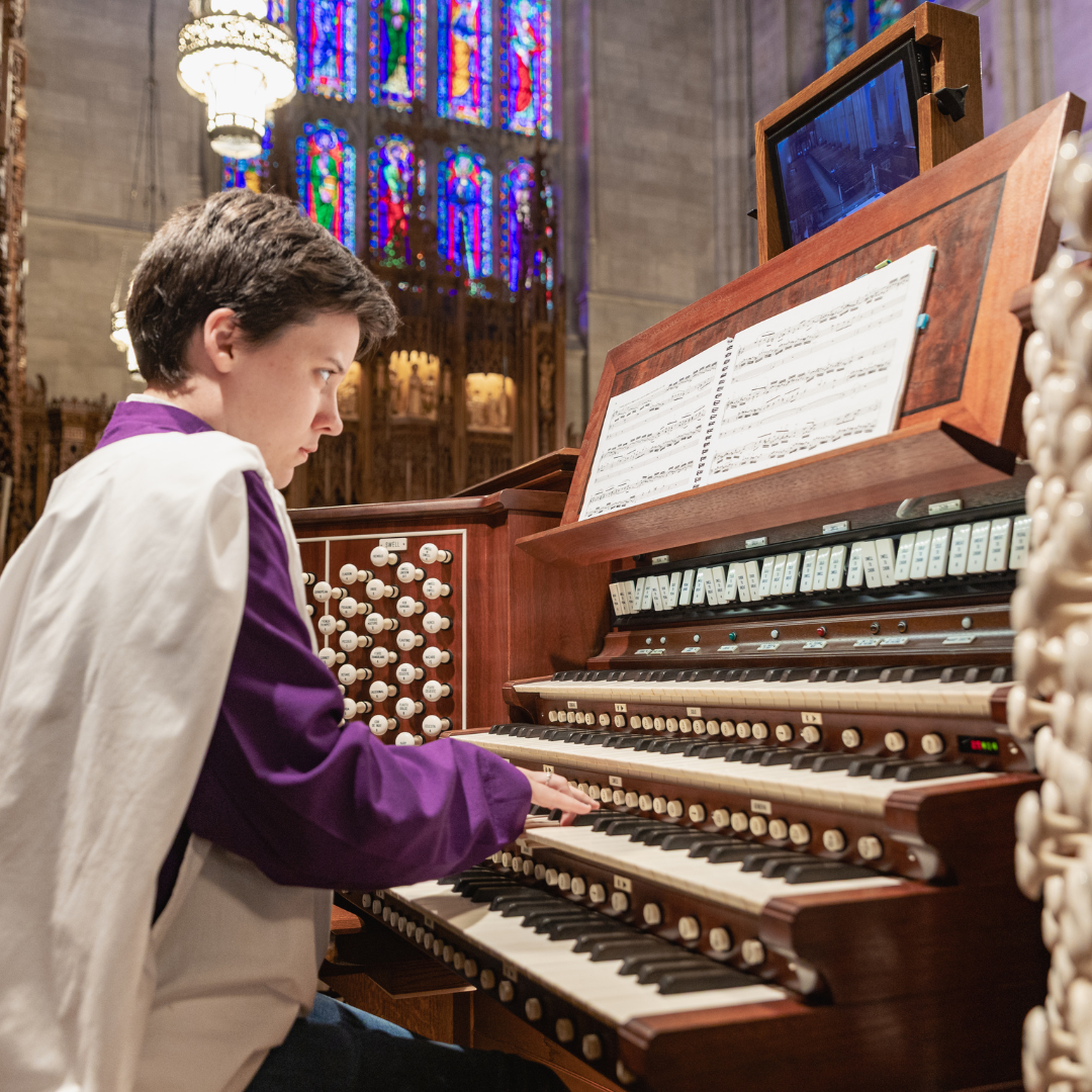 A musician in a purple and white robe plays a large pipe organ inside Duke Chapel. The scene shows multiple keyboards, rows of organ stops, and sheet music, with colorful stained-glass windows glowing in the background.