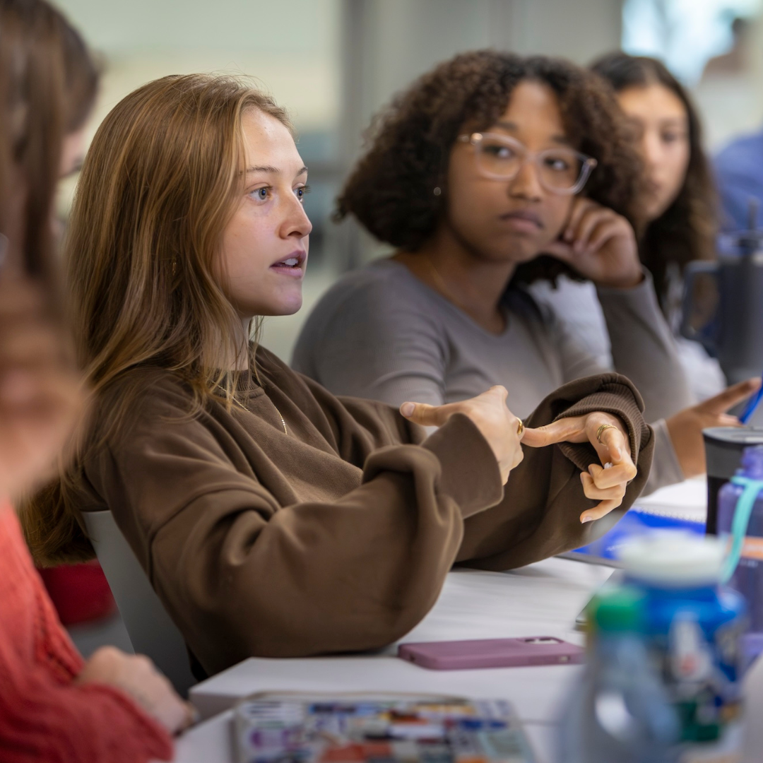 A student speaks during a classroom discussion, using hand gestures while others around the table listen attentively. Notebooks, water bottles, and name cards are spread across the table.