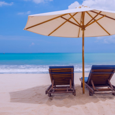 A photo of a beach. Blue skies and crystal clear blue sea crashing onto a sandy beach. There are 2 wooden deckchairs facing away from the camera, towards the sea, underneath a parasol.