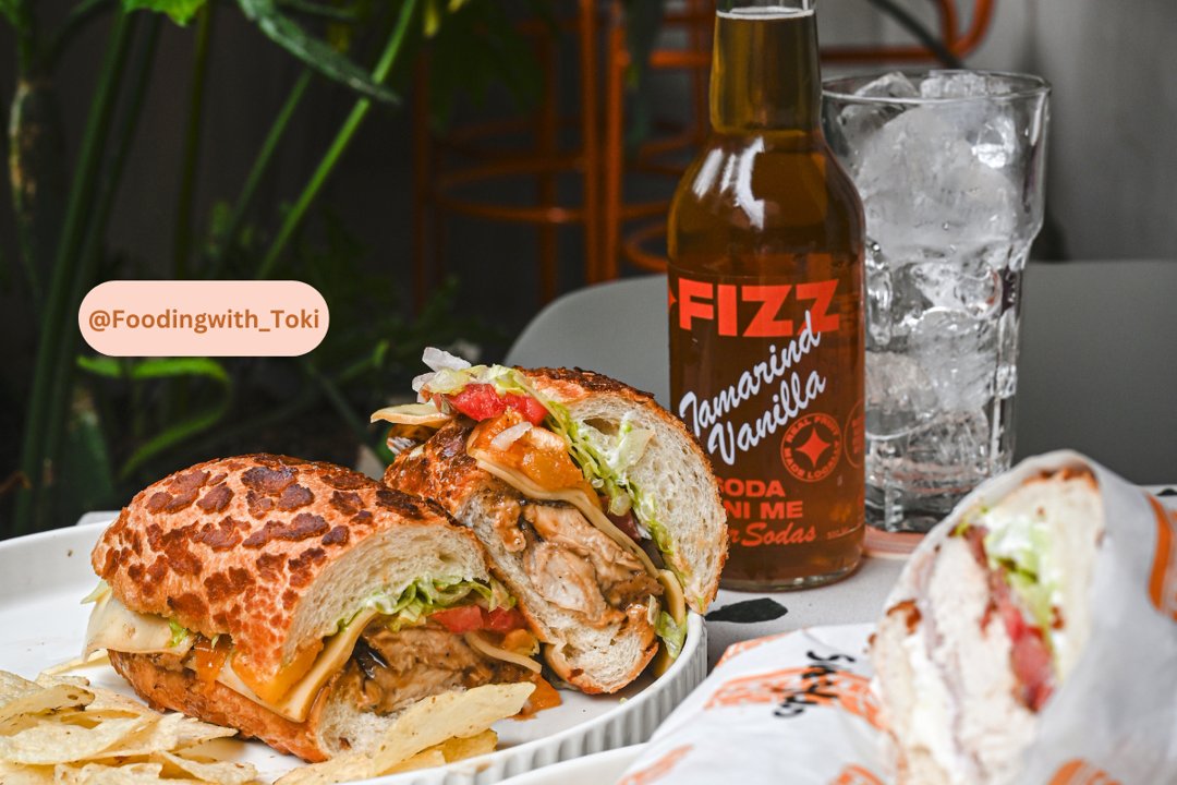A soda sits next to a Tiger Bread Sandwich full of meat and cheese